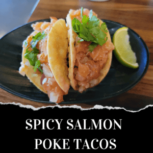 Two spicy salmon poke tacos topped with cilantro are served on a black plate with a lime wedge on the side.