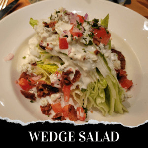 A wedge salad on a white plate topped with diced tomatoes, bacon pieces, blue cheese crumbles, chopped onions, and creamy dressing.