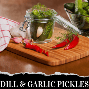 Glass jar of pickles with garlic, dill, and red chili peppers on a wooden cutting board, next to a bowl of cucumbers and a red and white cloth. Text reads "Dill & Garlic Pickles.