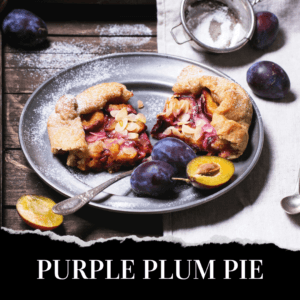 A rustic purple plum pie topped with sliced almonds is served on a gray plate with fresh plums and a spoon, set on a wooden table dusted with powdered sugar.