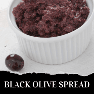 A white ramekin filled with black olive spread sits on a white surface beside a single whole black olive. Text below reads: “BLACK OLIVE SPREAD.”.