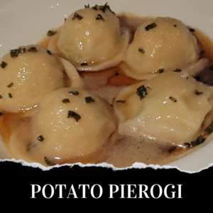 Five potato pierogi covered in a light brown sauce and garnished with chopped herbs, arranged on a white plate.