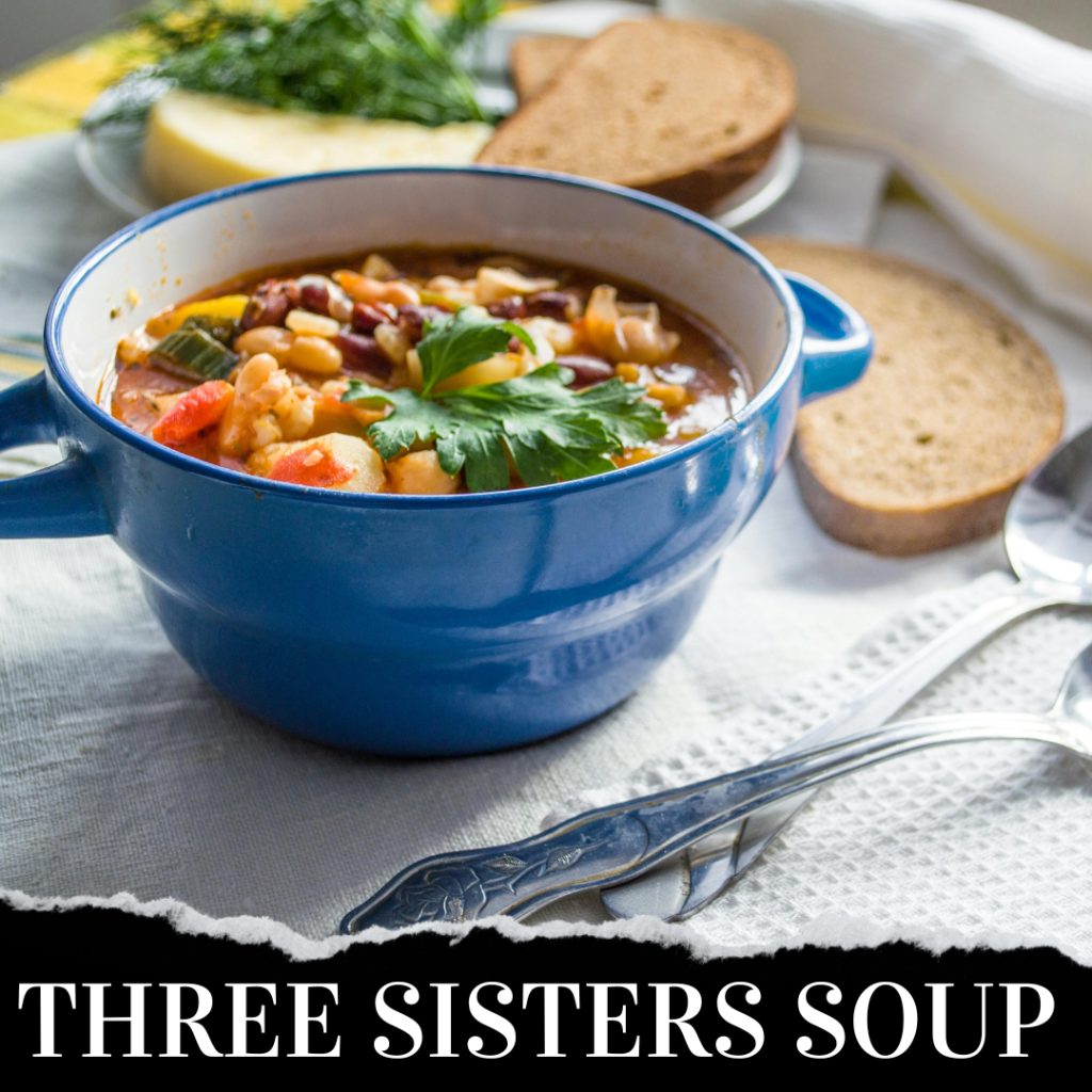 A blue bowl of Three Sisters Soup garnished with herbs. Two slices of brown bread, a yellow vegetable, and herbs in the background on a white cloth. Two spoons are beside the bowl.