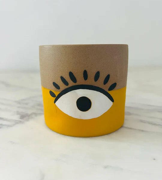 A small cylindrical pot with a beige and yellow design featuring a stylized eye in the middle.