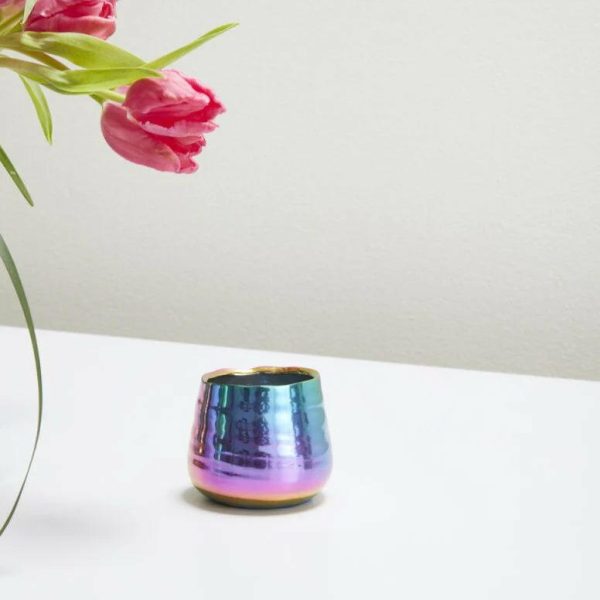 A small iridescent candle holder sits on a reflective surface next to partially visible pink tulips.