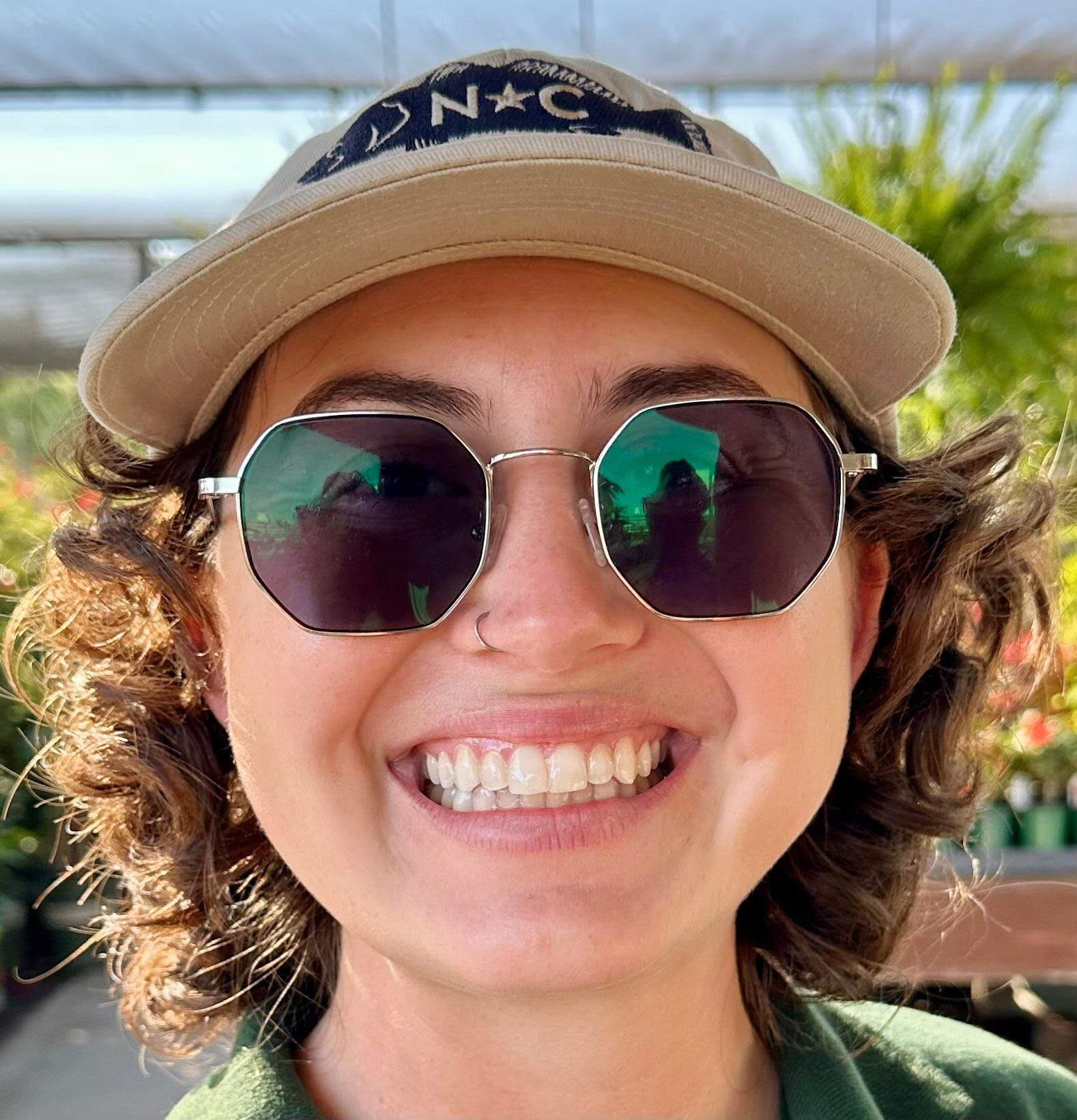 A person with short curly hair, wearing a beige cap and reflective sunglasses, smiles outdoors in a sunny, leafy environment.